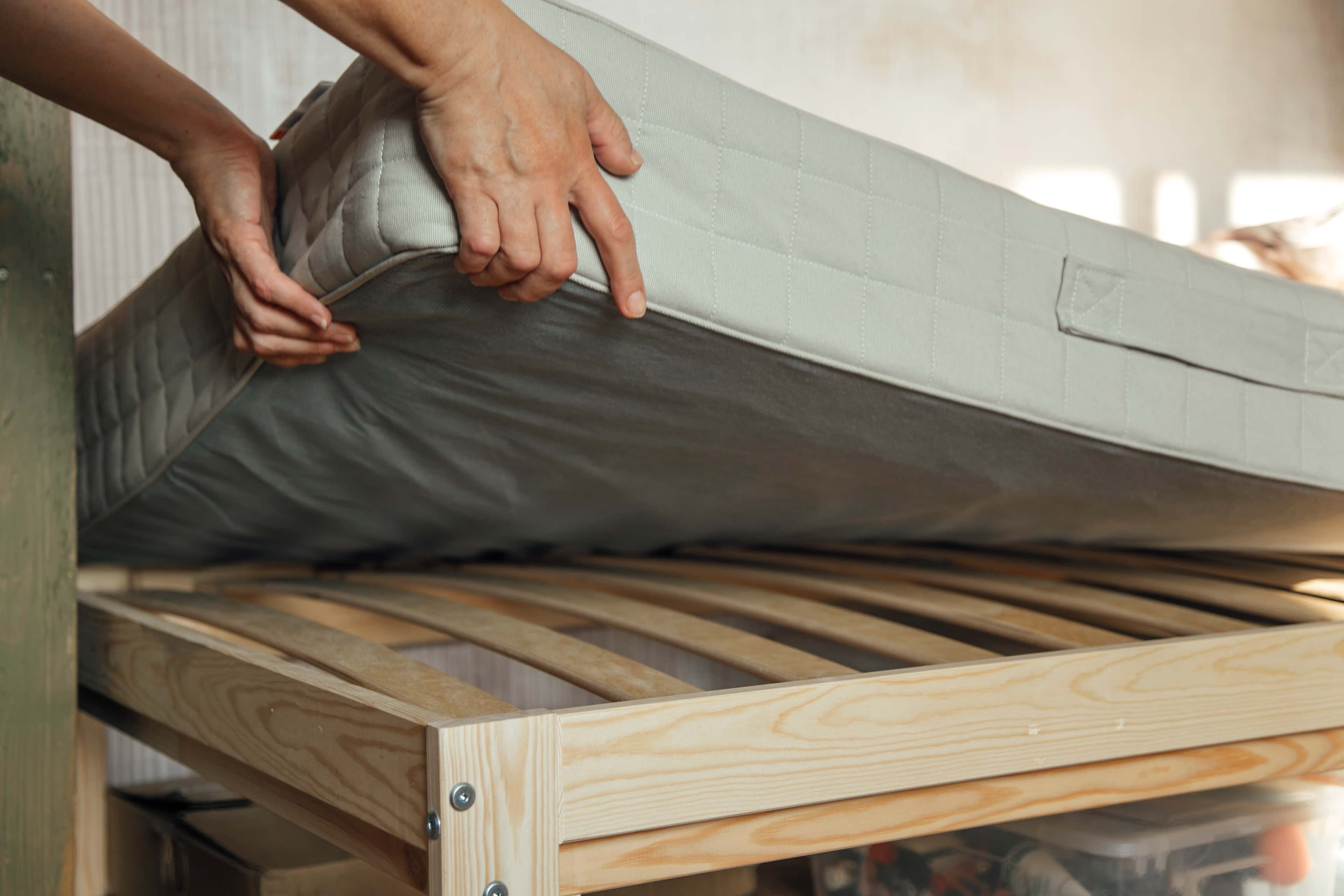 Hands lifting a mattress to inspect a wooden bed frame.