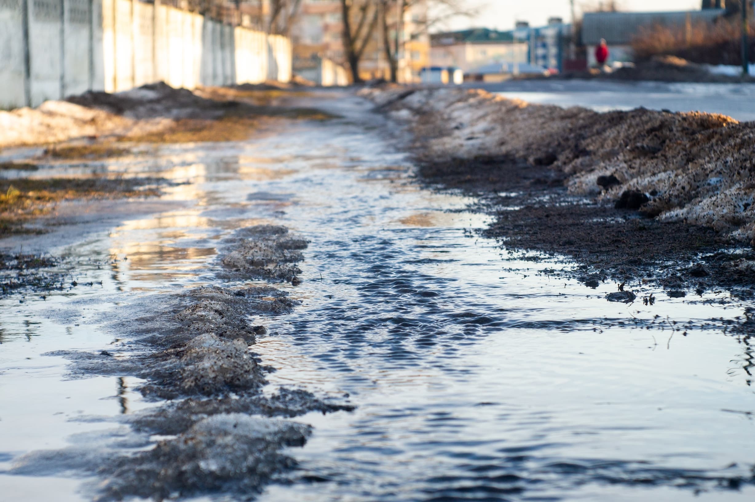 Urban street with water accumulation and erosion near a concrete fence.