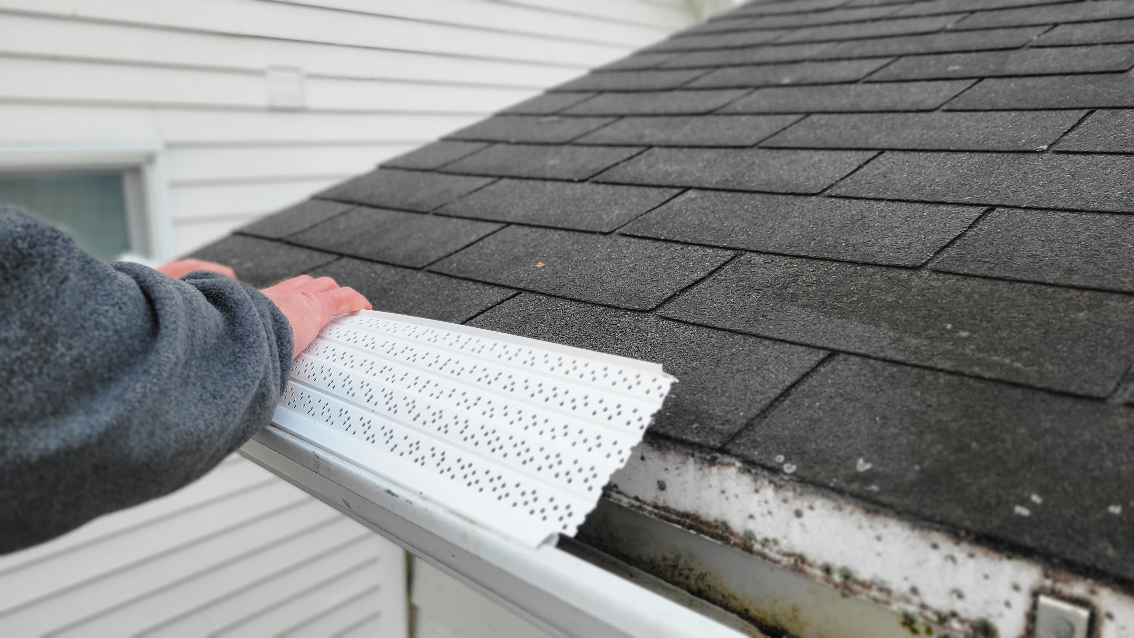 Person installing eavestrough guards on a roof with dark shingles.