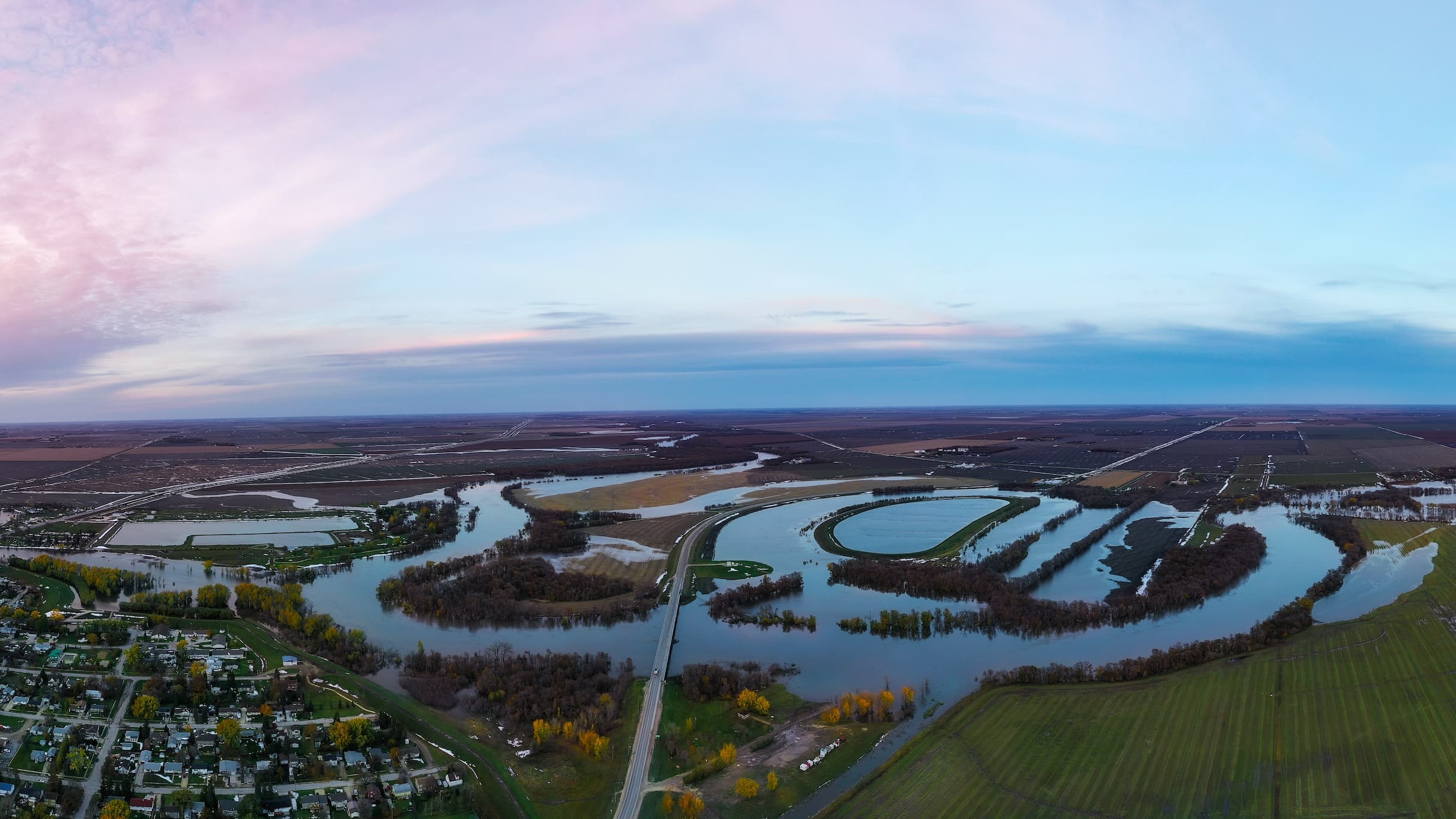 Aerial view of flooded farmland and residential areas under a pink and blue sky.