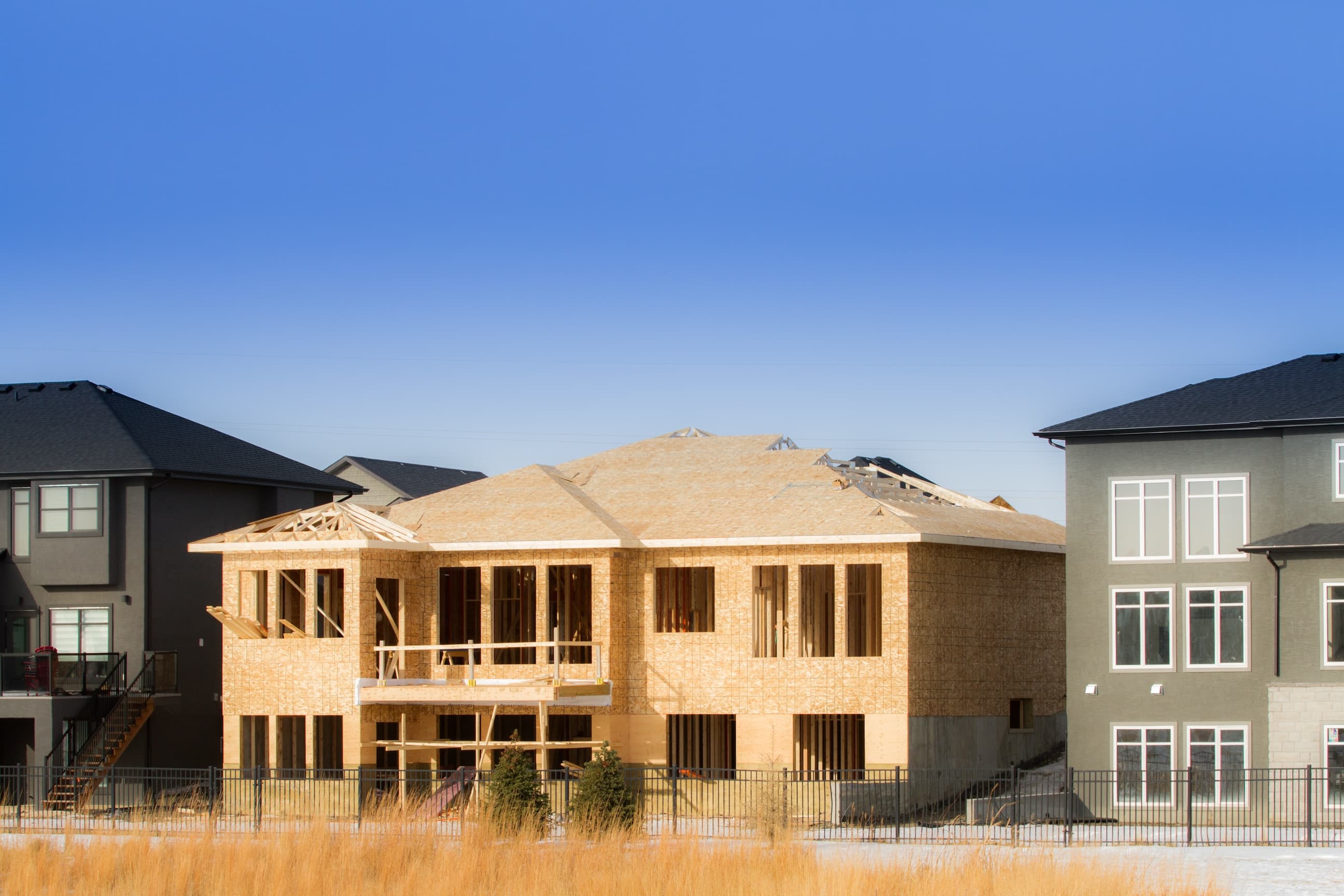 Partially constructed home between two completed houses in a suburban area under a blue sky.