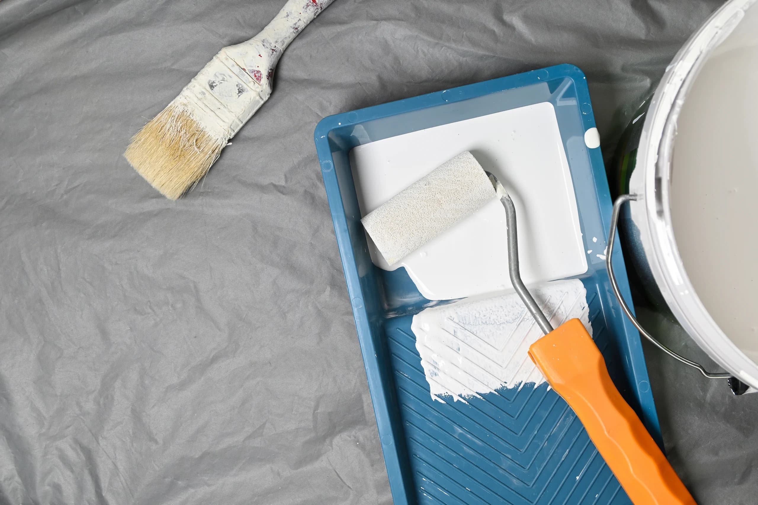 Painting tools on drop cloth with brush, roller, tray, and paint can.