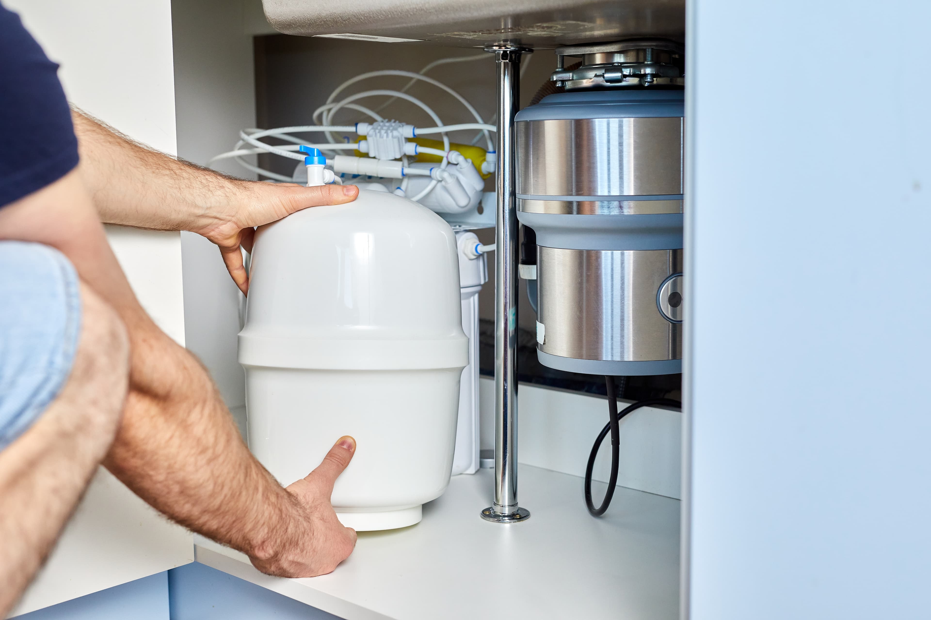Person installing reverse osmosis tank under kitchen sink with pipes and metal filter.