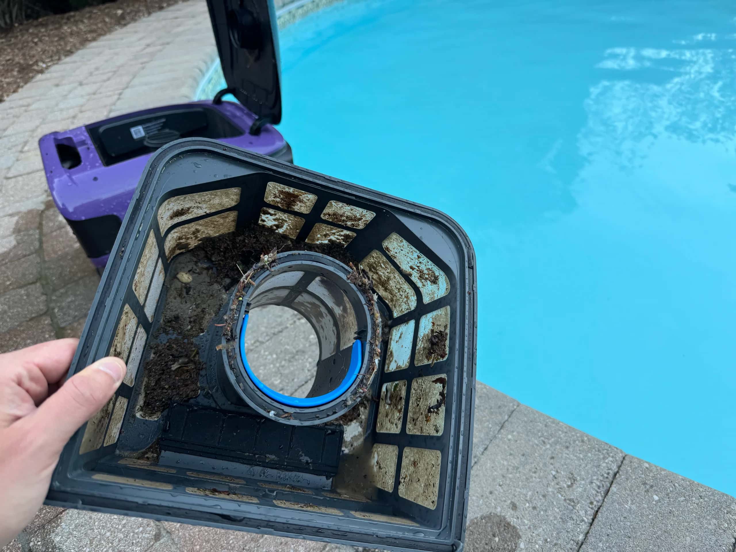 Robotic pool cleaner filter with debris, purple cleaner open on deck beside pool.