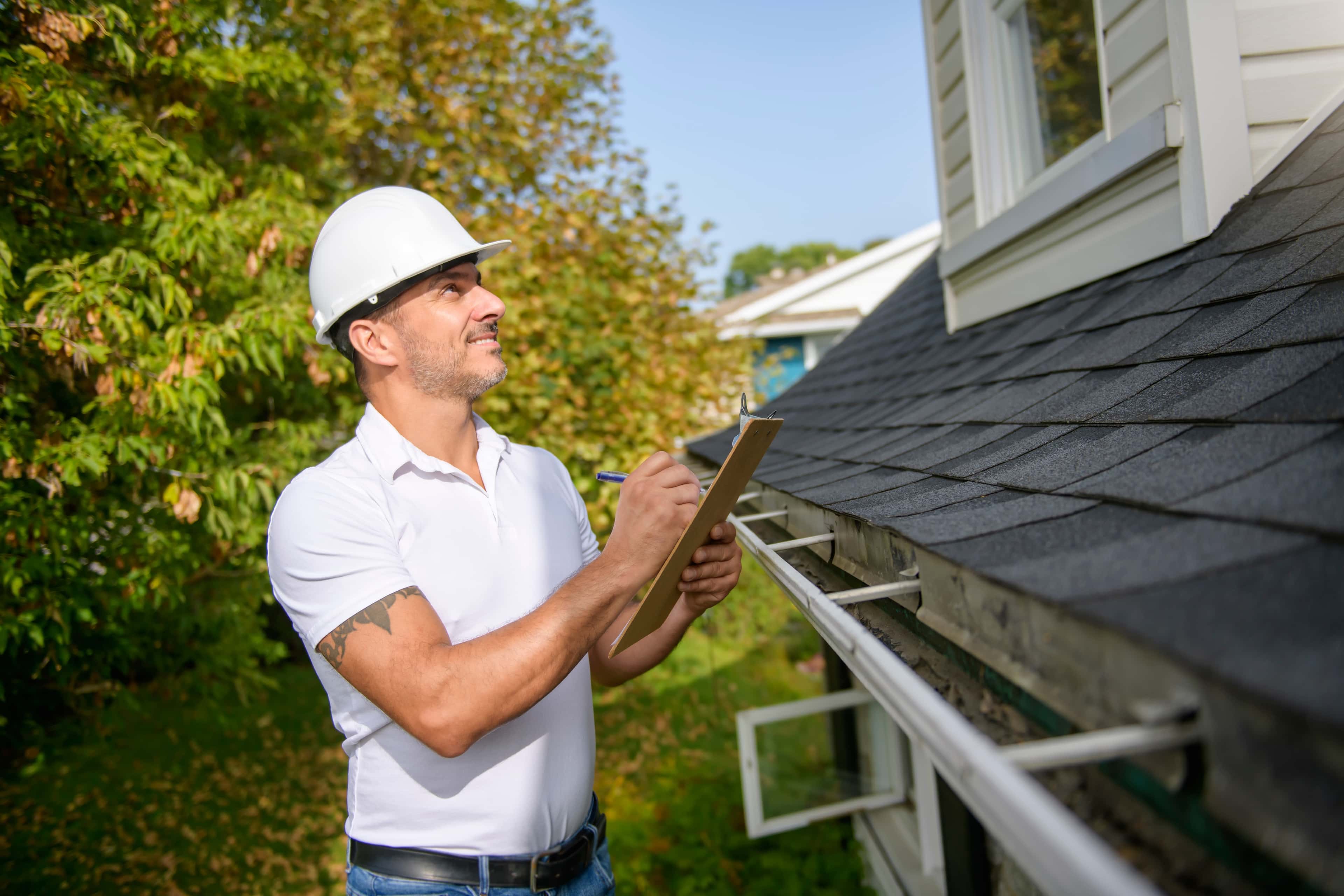 Contractor inspecting roof shingles, taking notes on a clipboard on a sunny day.